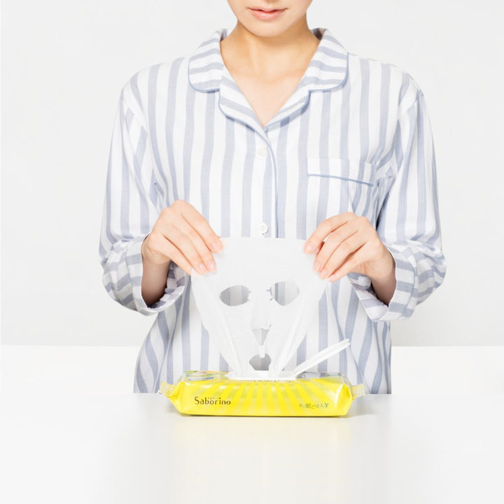 Person i randiga pyjamas håller upp ett Saborino Morning Facial Sheet Mask N 32st ovanför en öppen förpackning.
