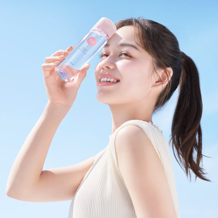 Leende kvinna utomhus håller momopuri Moist Barrier Lotion Regular 200ml nära ansiktet mot blå himmel.